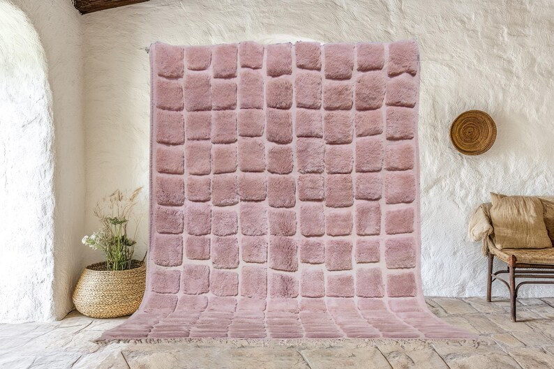 May include: A large, rectangular, blush-colored rug with a textured, square pattern. The rug has a soft, plush appearance and a fringed edge. The rug is displayed indoors, leaning against a textured white wall.