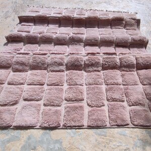May include: A dusty rose-colored rug with a textured, tufted design. The rug features a repeating pattern of rectangular blocks, creating a plush, dimensional surface. The rug's edges are finished with a simple border and fringe.