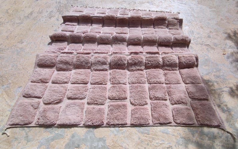 May include: A dusty rose-colored rug with a textured, tufted design. The rug features a repeating pattern of rectangular blocks, creating a plush, dimensional surface. The rug's edges are finished with a simple border and fringe.