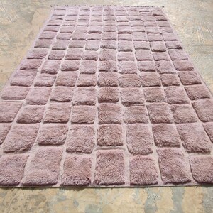 May include: A rectangular rug in a muted mauve color, featuring a repeating pattern of raised, square tufts. The rug has a soft, textured appearance and is placed on a light-colored surface.