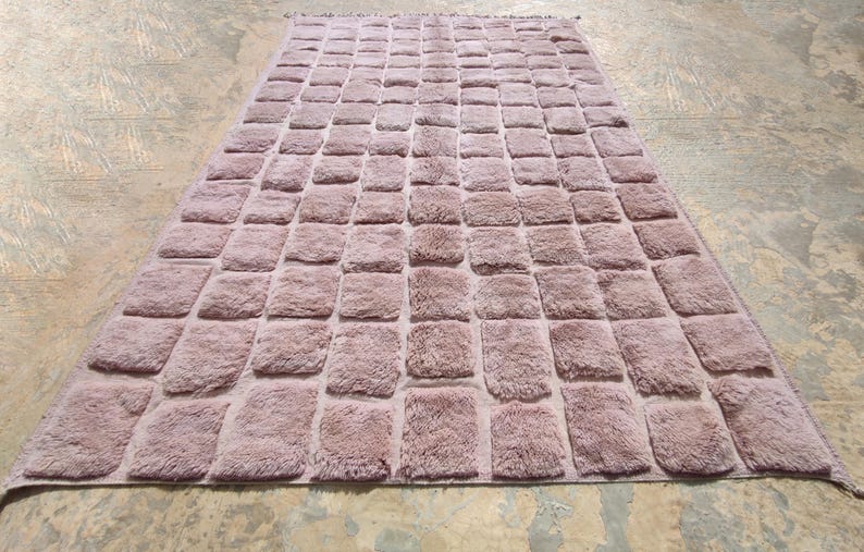May include: A rectangular rug in a muted mauve color, featuring a repeating pattern of raised, square tufts. The rug has a soft, textured appearance and is placed on a light-colored surface.