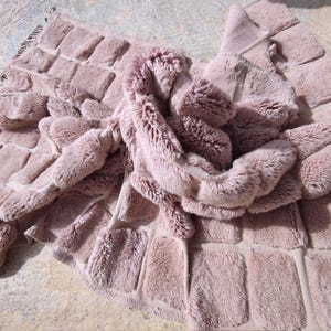 May include: A plush, dusty rose-colored rug with a textured, rectangular pattern. The rug has a soft, furry appearance and is folded over itself, showcasing its luxurious texture. The edges are finished with a fringe.