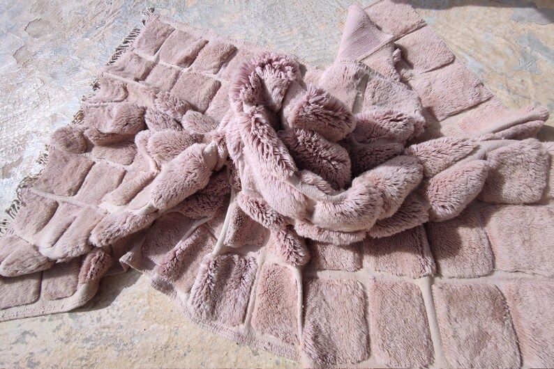 May include: A plush, dusty rose-colored rug with a textured, rectangular pattern. The rug has a soft, furry appearance and is folded over itself, showcasing its luxurious texture. The edges are finished with a fringe.