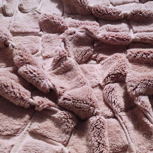 May include: Close-up of a plush, textured blanket in a dusty rose color. The blanket features a woven design with raised, rectangular sections, creating a soft, inviting appearance. The surface has a furry texture, suggesting warmth and comfort.