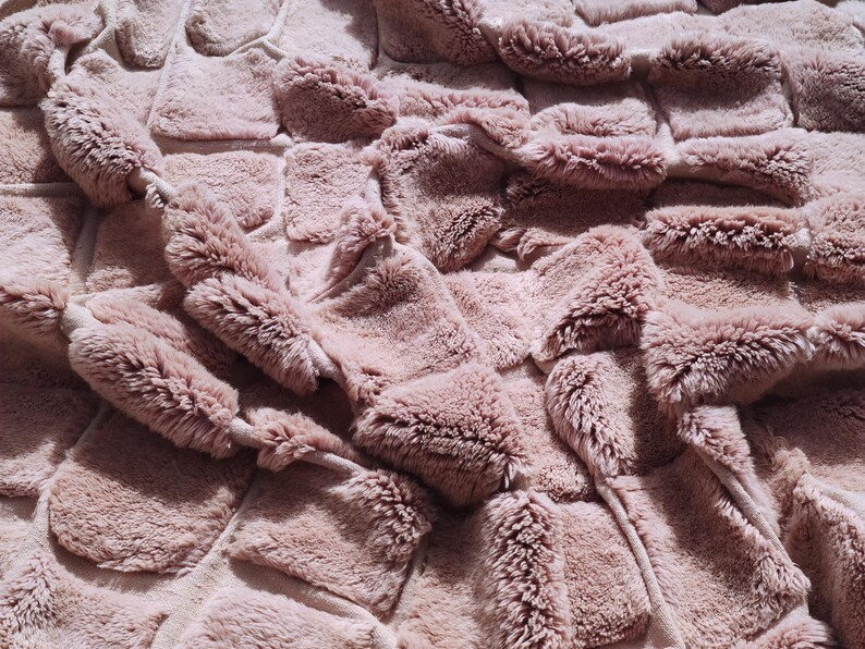 May include: Close-up of a plush, textured blanket in a dusty rose color. The blanket features a woven design with raised, rectangular sections, creating a soft, inviting appearance. The surface has a furry texture, suggesting warmth and comfort.