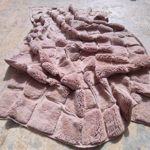 May include: A dusty rose-colored, faux fur throw blanket. The blanket features a patchwork design with rectangular blocks of plush fur. The edges are finished with a simple border.