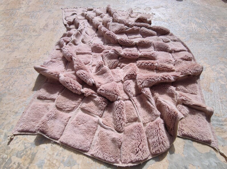 May include: A dusty rose-colored, faux fur throw blanket. The blanket features a patchwork design with rectangular blocks of plush fur. The edges are finished with a simple border.