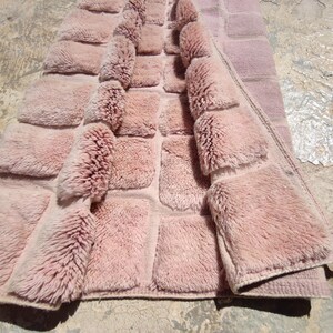 May include: A dusty rose-colored rug with a textured, tufted design. The rug features a grid pattern with raised, fluffy squares, creating a plush, inviting appearance. The edges are neatly finished, and the rug is partially folded, revealing its construction.
