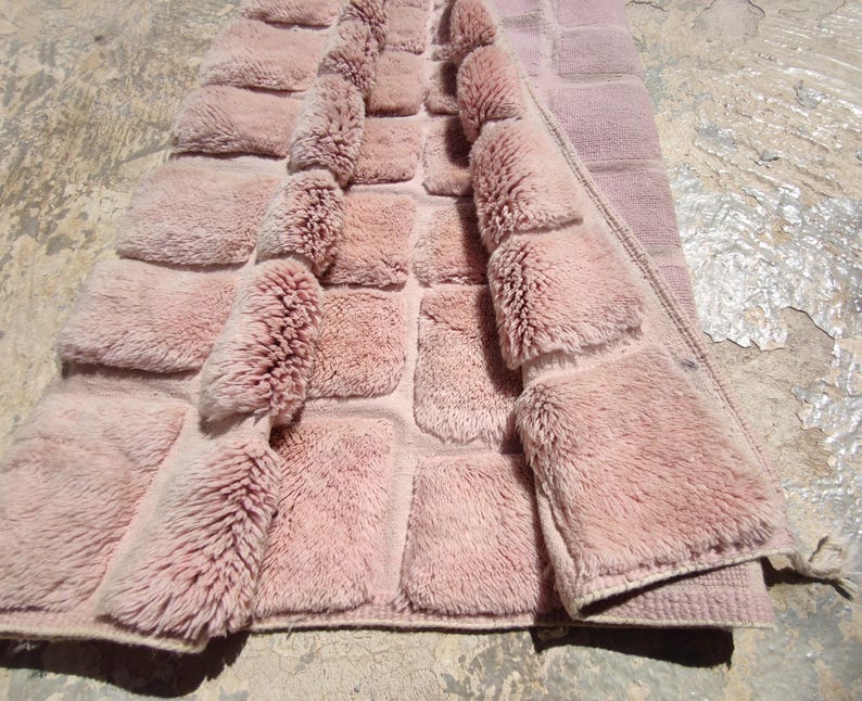 May include: A dusty rose-colored rug with a textured, tufted design. The rug features a grid pattern with raised, fluffy squares, creating a plush, inviting appearance. The edges are neatly finished, and the rug is partially folded, revealing its construction.