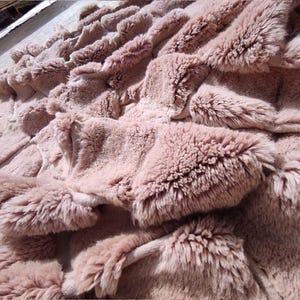 May include: Close-up of a plush, textured blanket in a muted rose color. The blanket features a layered design with raised, fluffy sections, creating a soft, inviting appearance. The overall aesthetic suggests comfort and warmth.
