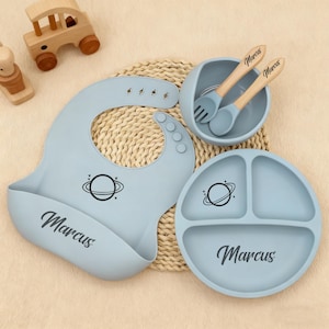 May include: A baby feeding set in light blue, including a bib, divided plate, bowl, and spoon and fork. Each piece has a planet graphic and the name "Marcus". The spoon and fork have wooden handles. Wooden toy car and figure are in the background.