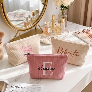 Può includere: Tre pochette in velluto a coste color crema e rosa. La pochette color crema a sinistra presenta il nome Clara in rosa con un fiocco. La pochette color crema a destra presenta il nome Roberta in marrone. La pochette rosa davanti ha il nome Eleanor.