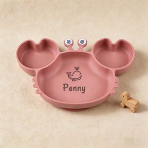 May include: A dusty rose silicone divided plate shaped like a crab with two eye stalks. The centre features a black line drawing of a whale above the name Penny in a dark font. A small wooden toy animal on wheels sits to the right of the plate.