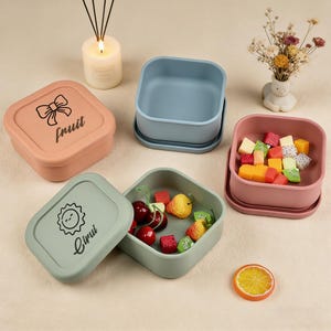 Puede incluir: Una colección de recipientes cuadrados de comida coloridos. Uno es melocotón con un lazo y la palabra "fruit", otro es verde con un sol y la palabra "Cirui". Los recipientes están llenos de fruta. También hay una vela y un pequeño jarrón.