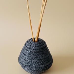 May include: A dark blue woven vase with a round base and a narrow opening, containing several light brown wooden reeds. The vase has a textured, layered design. The background is a solid, light beige colour.