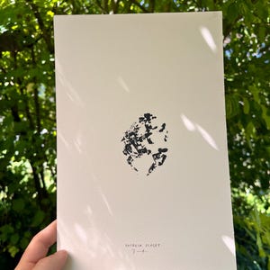 Könnte beinhalten: Ein cremefarbenes Kunstwerk mit einem zentralen abstrakten schwarzen Tintenklecks. Der Name des Künstlers, "PATRICIA SCALET", und eine Signatur sind unten mit schwarzer Tinte geschrieben. Das Kunstwerk wird vor einem Hintergrund aus grünem Laub gehalten.