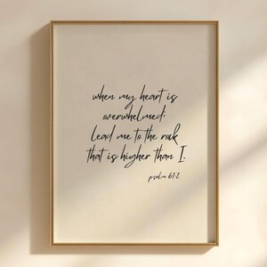 Può includere: Una stampa artistica da parete rettangolare in una sottile cornice dorata con testo nero scritto a mano su uno sfondo beige chiaro. Il testo recita: When my heart is overwhelmed; lead me to the rock that is higher than I. Psalm 61:2.