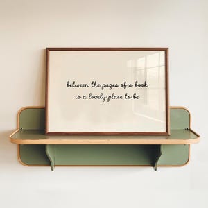 May include: A rectangular wall shelf with a sage green finish and light wood trim, holding a framed print. The print features the black text: between the pages of a book is a lovely place to be, centred on a cream-coloured background.