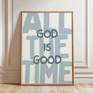 Pu&ograve; includere: Una stampa artistica rettangolare incorniciata con uno sfondo color crema chiaro e grandi lettere a blocchi azzurre che compongono il testo ALL THE TIME. Centrata sopra il testo blu, la frase GOD IS GOOD &egrave; scritta in lettere grigio antracite.