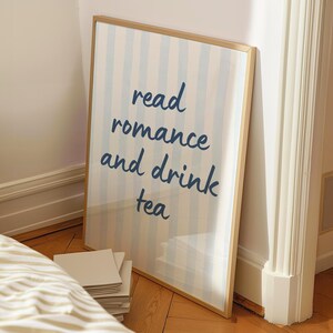 May include: A rectangular wall art print in a thin gold frame featuring vertical light blue and white stripes. The text read romance and drink tea is printed in a dark blue, handwritten-style font centered on the striped background.