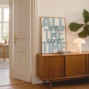 Pu&ograve; includere: Una stampa artistica rettangolare incorniciata con il testo ALL GOD IS GOOD TIME in lettere blu scuro sovrapposte a grandi lettere in stampatello blu chiaro su sfondo bianco. La stampa &egrave; esposta su una credenza in legno accanto a una lampada.