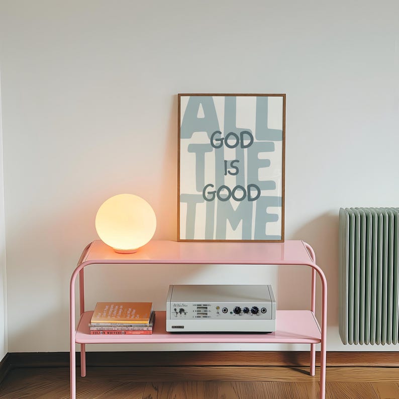 Pu&ograve; includere: Una stampa da parete rettangolare incorniciata con il testo ALL GOD IS GOOD TIME in lettere grigio scuro su sfondo azzurro. La stampa poggia su una console in metallo rosa accanto a una lampada sferica bianca, sopra libri e un dispositivo elettronico grigio.