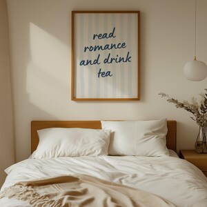 May include: A rectangular wall art print in a wooden frame featuring vertical light blue stripes and the dark blue text "read romance and drink tea" centered on a white background, displayed above a bed with white bedding and pillows.