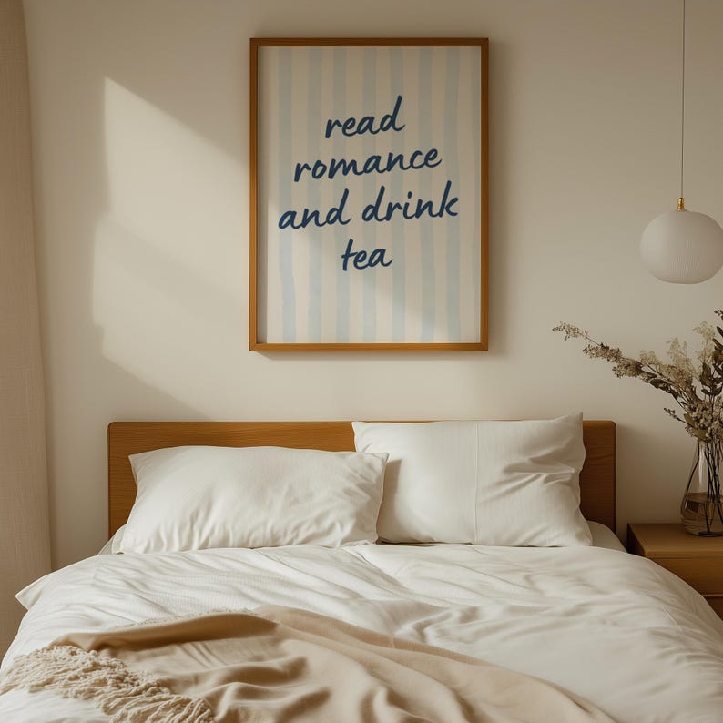 May include: A rectangular wall art print in a wooden frame featuring vertical light blue stripes and the dark blue text "read romance and drink tea" centered on a white background, displayed above a bed with white bedding and pillows.