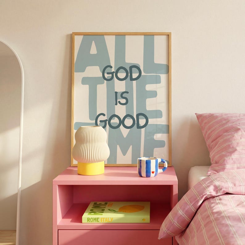 Pu&ograve; includere: Una stampa rettangolare in una cornice di legno chiaro con il testo ALL GOD IS GOOD TIME in lettere blu scuro su sfondo azzurro. Sotto, una lampada color crema a coste con base gialla e una tazza a righe blu e bianche su un comodino rosa.