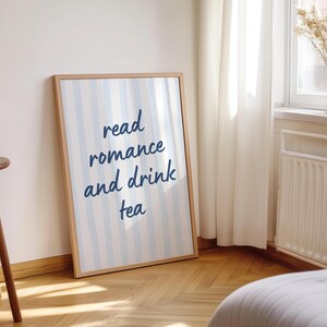 May include: A rectangular framed wall art print featuring vertical light blue and white stripes. The center of the print displays the dark blue text read romance and drink tea in a casual, handwritten script font. The frame is a light wood tone.
