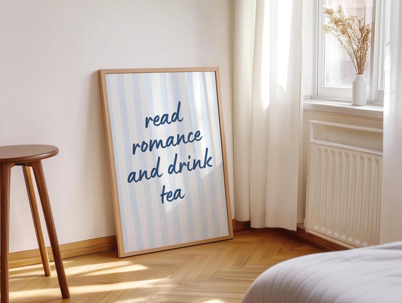 May include: A rectangular framed wall art print featuring vertical light blue and white stripes. The center of the print displays the dark blue text read romance and drink tea in a casual, handwritten script font. The frame is a light wood tone.