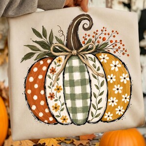 May include: A beige sweatshirt featuring a large graphic of a pumpkin with vertical segments. The pumpkin has a brown stem, a tan bow, and patterns including white polka dots, orange flowers, green gingham, and white daisies with orange berries.