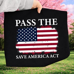 Peut inclure: Sac fourre-tout noir avec le drapeau américain et le texte "PASS THE" en haut et "SAVE AMERICA ACT" en bas. Le drapeau présente des rayures rouges et blanches et un champ bleu avec des étoiles blanches. Le sac est tenu devant un fond de fleurs roses et d'herbe verte.