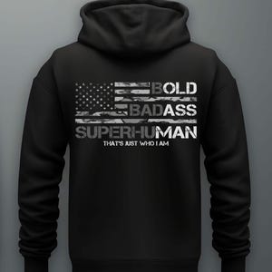 Puede incluir: Sudadera con capucha negra con un diseño de bandera estadounidense desgastada. Las palabras "BOLD BADASS SUPERHUMAN" están impresas en la bandera, con la frase "THAT'S JUST WHO I AM" debajo.