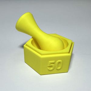 Puede incluir: Un juego de mortero y maja de color amarillo brillante. El mortero tiene forma hexagonal con el número "50" en relieve en un lado. La maja tiene un mango redondeado y un extremo de molienda ensanchado. El juego es de material mate.