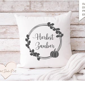 Puede incluir: Un cojín cuadrado blanco con un diseño de corona negra, el texto alemán "Herbst Zauber" y una ilustración de calabaza. El cojín está sobre una superficie blanca con un fondo de madera y una manta de punto blanca.