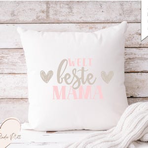 May include: A white square cushion with the German text "WELT beste MAMA" in grey and pink, and two grey hearts. The cushion is on a light wood surface. A white knitted blanket is in the bottom right corner.