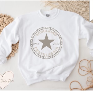 Könnte beinhalten: Weißes Sweatshirt mit Rundhalsausschnitt und einem kreisförmigen Grafik in Taupe. Die Grafik zeigt einen Stern und die Worte "SCHULKIND ERSTKLASSIG since 2026". Das Sweatshirt liegt auf einer gewebten Matte.