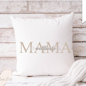 May include: A white square cushion with the words "MAMA" and "weltbeste" in light brown. The cushion is on a wooden surface with a white knitted blanket. A heart-shaped wooden cutout is in the lower left corner.