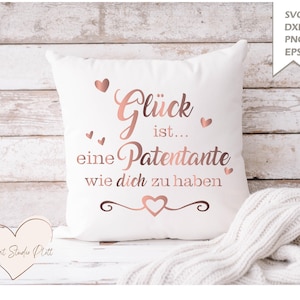 May include: White square pillow with the German text "Glück ist... eine Patentante wie dich zu haben" in rose gold script and heart accents. A heart-shaped wooden cutout and a white knitted blanket are also visible.