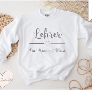 May include: White crewneck sweatshirt with the word "Lehrer" in gray script above a horizontal line with a heart, and the text "Ein Mann mit Klasse" below. The sweatshirt is displayed on a neutral background with decorative elements.