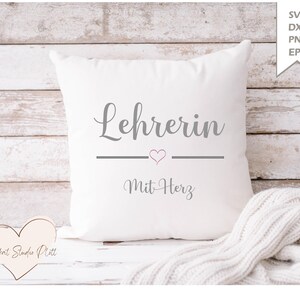 May include: A white square pillow with the word "Lehrerin" in gray script, a gray line with a pink heart, and the words "Mit Herz" in gray script. A white knitted blanket is draped on the bottom right. The background is a white wooden surface.