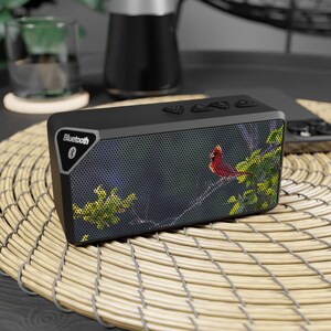 May include: Black rectangular Bluetooth speaker with a nature scene featuring a red cardinal bird perched on a branch. The speaker has control buttons on top and a Bluetooth logo. It sits on a woven mat.