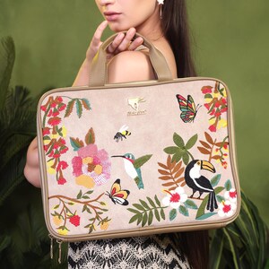 May include: Beige laptop bag with embroidered floral and wildlife designs. The bag features a hummingbird, toucan, butterflies, and a bee. The bag has a zipper closure and a handle.