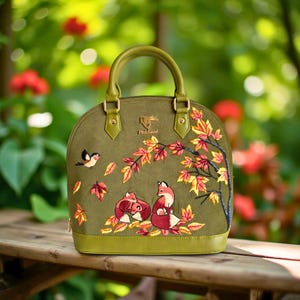 Peut inclure: Sac à main vert olive en forme de dôme avec une scène d'automne brodée. Il présente deux renards, un oiseau et des feuilles colorées. Le sac a une poignée supérieure et des ferrures dorées.