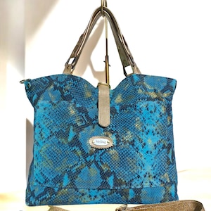 Peut inclure: Sac fourre-tout turquoise et or à motif peau de serpent avec une bandoulière assortie. Le sac a une forme rectangulaire avec un dessus incurvé et une petite étiquette qui dit "Melibag". Les poignées et la bandoulière sont de couleur or métallique.