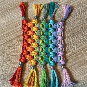 Könnte beinhalten: Ein handgefertigter Makramee-Untersetzer in Regenbogenfarben. Er zeigt Rot, Orange, Gelb, Grün, Blau und Lila. Jeder Quasten-Abschluss ist mit einem silbernen Band versehen. Der Untersetzer ist aus Baumwollschnur gefertigt.