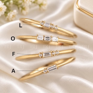 May include: Four gold rings arranged vertically, each featuring a unique combination of baguette and round-cut clear stones. The rings are labeled with the letters L, O, F, and A, and the background features the repeating text TRJ.