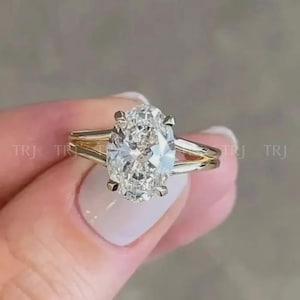 Könnte beinhalten: Ein goldener Verlobungsring mit einem großen, oval geschliffenen Diamanten. Der Ring hat ein geteiltes Schaftdesign, wobei sich das Band auf beiden Seiten des Diamanten in zwei separate Stränge aufteilt. Der Diamant wird von vier Krappen gehalten.