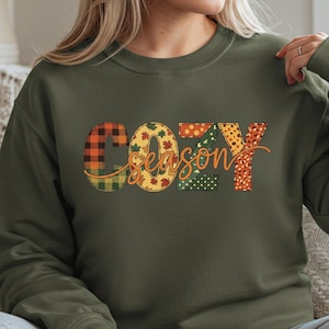 May include: Military green sweatshirt with the words "COZY SEASON" in a colorful, patchwork design. The letters are filled with plaid, polka dots, and leaf patterns. The sweatshirt is a long-sleeved crew neck.